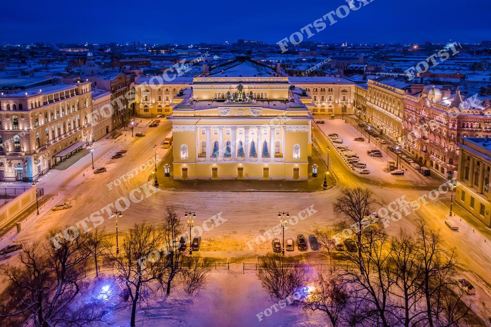 На фотографии изображен Александринский театр в зимний вечер, окруженный величественными зданиями на площади Островского. Фасад театра украшен иллюминацией и рождественскими декорациями, придавая праздничное настроение. Зимний пейзаж города с подсвеченными зданиями дополняет волшебную атмосферу центральных улиц Петербурга. Александринский театр является одним из символов русской театральной культуры.
